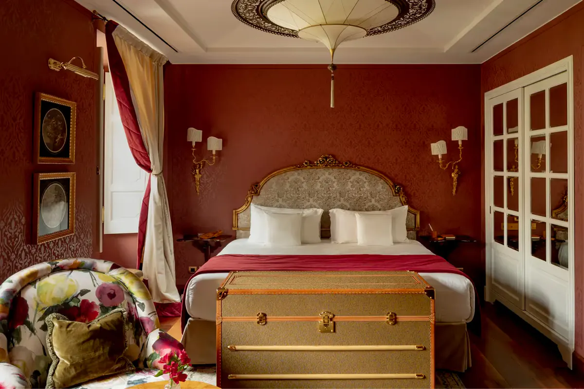 A bedroom with deep red walls, gilded bed, and tall white doors opening to windows