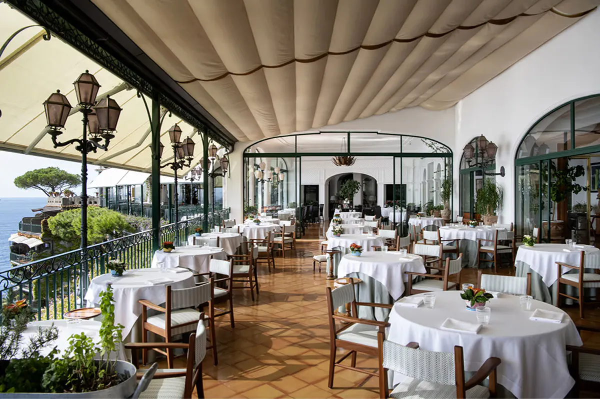 Covered terrace dining area with round white-clothed tables, wooden chairs, sea views, Italian coastal setting