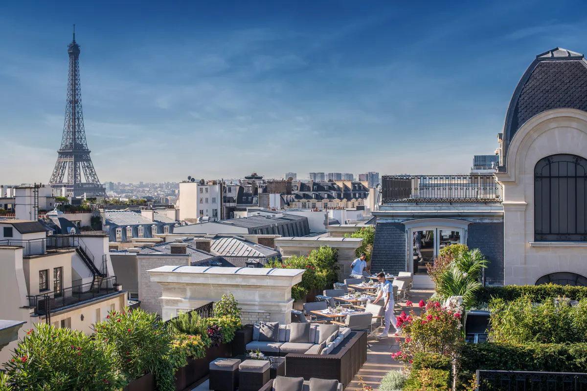 Rooftop terrace with white stone columns, potted plants, and dining tables overlooking Paris