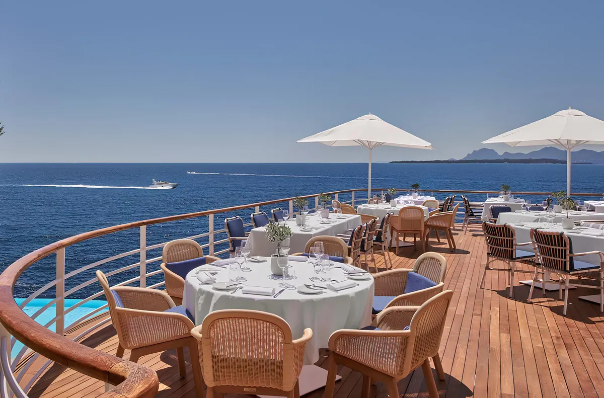 A terrace restaurant with round tables, wooden chairs, white umbrellas, and sea views