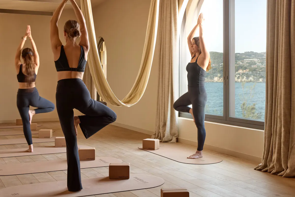 Guests hold a yoga pose on cork mats before arched windows overlooking the sea
