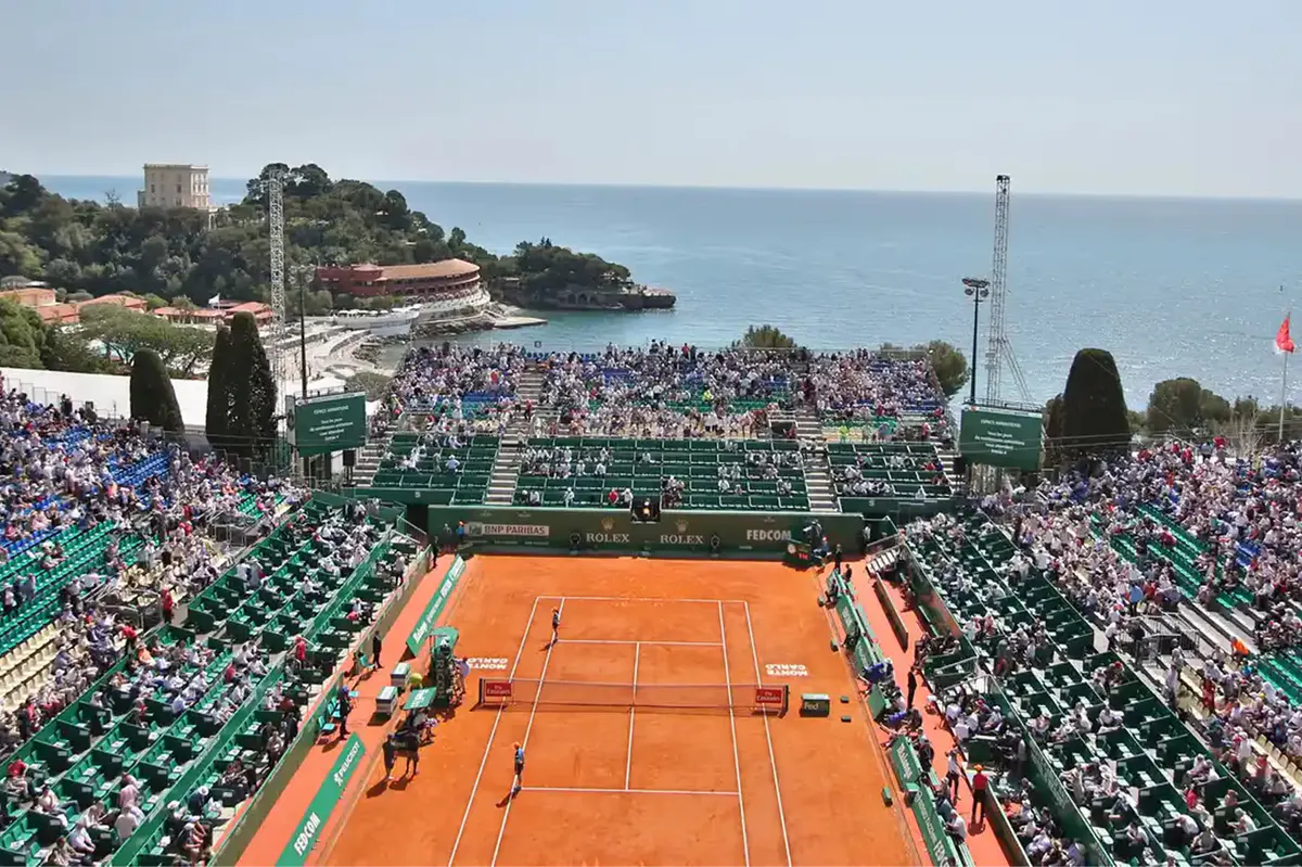 Red clay tennis court with spectator stands overlooking Mediterranean shoreline