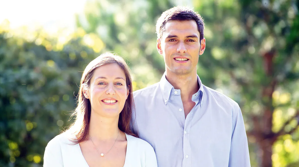 Two people smiling outdoors with green foliage in background