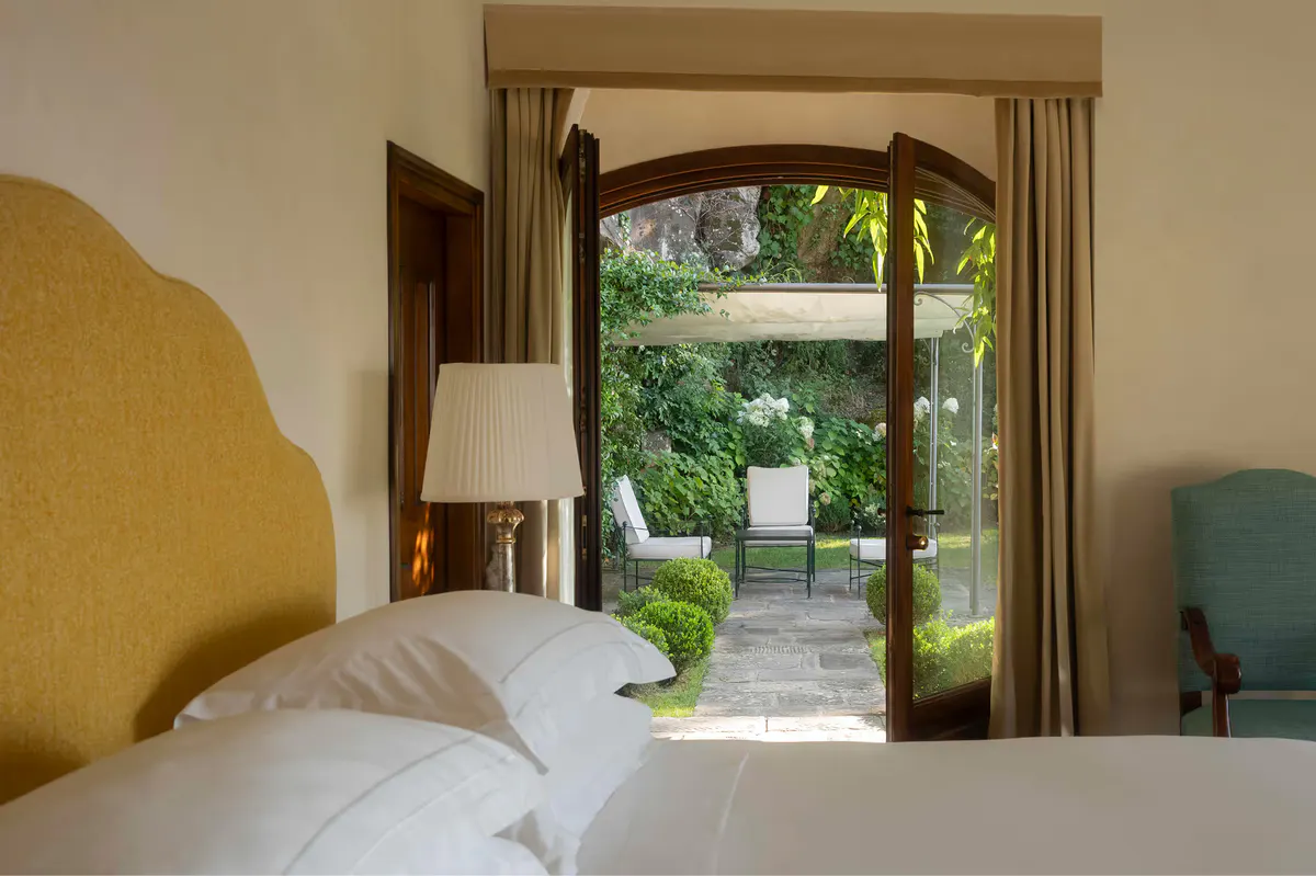 A bright bedroom with arched doorway opening to a garden view, mustard accent chair, and terracotta floor