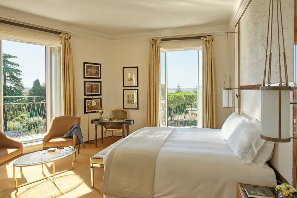 A guest room with large windows, a bed with beige linens, wooden furniture, and views of Rome’s gardens