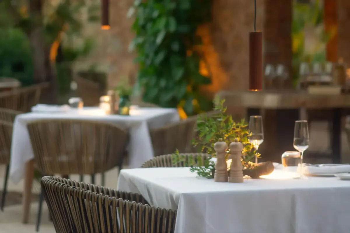 Two restaurant tables with white linens in a courtyard with stone walls and lush greenery