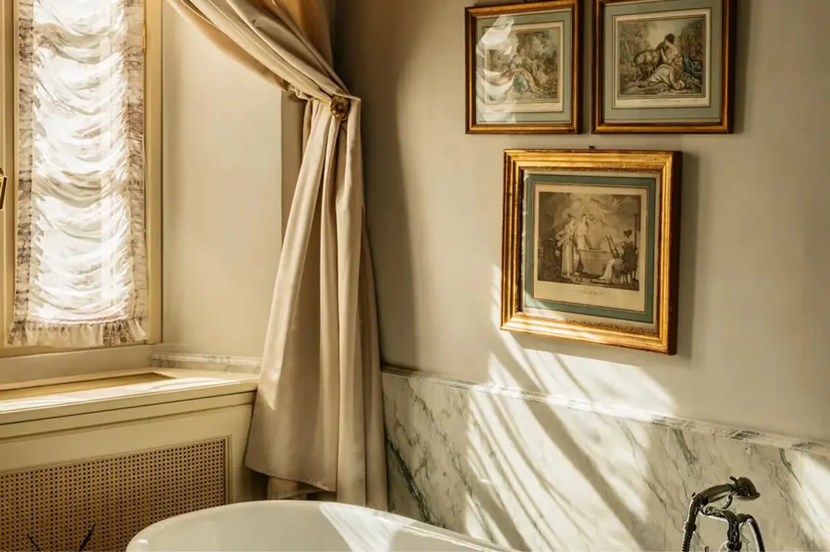 A sun-filled bedroom corner with framed artwork, window, and cream bedding