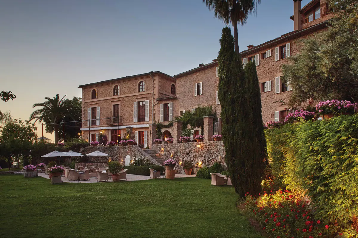 Estate buildings in evening light with manicured grounds, cypress trees, and palm tree