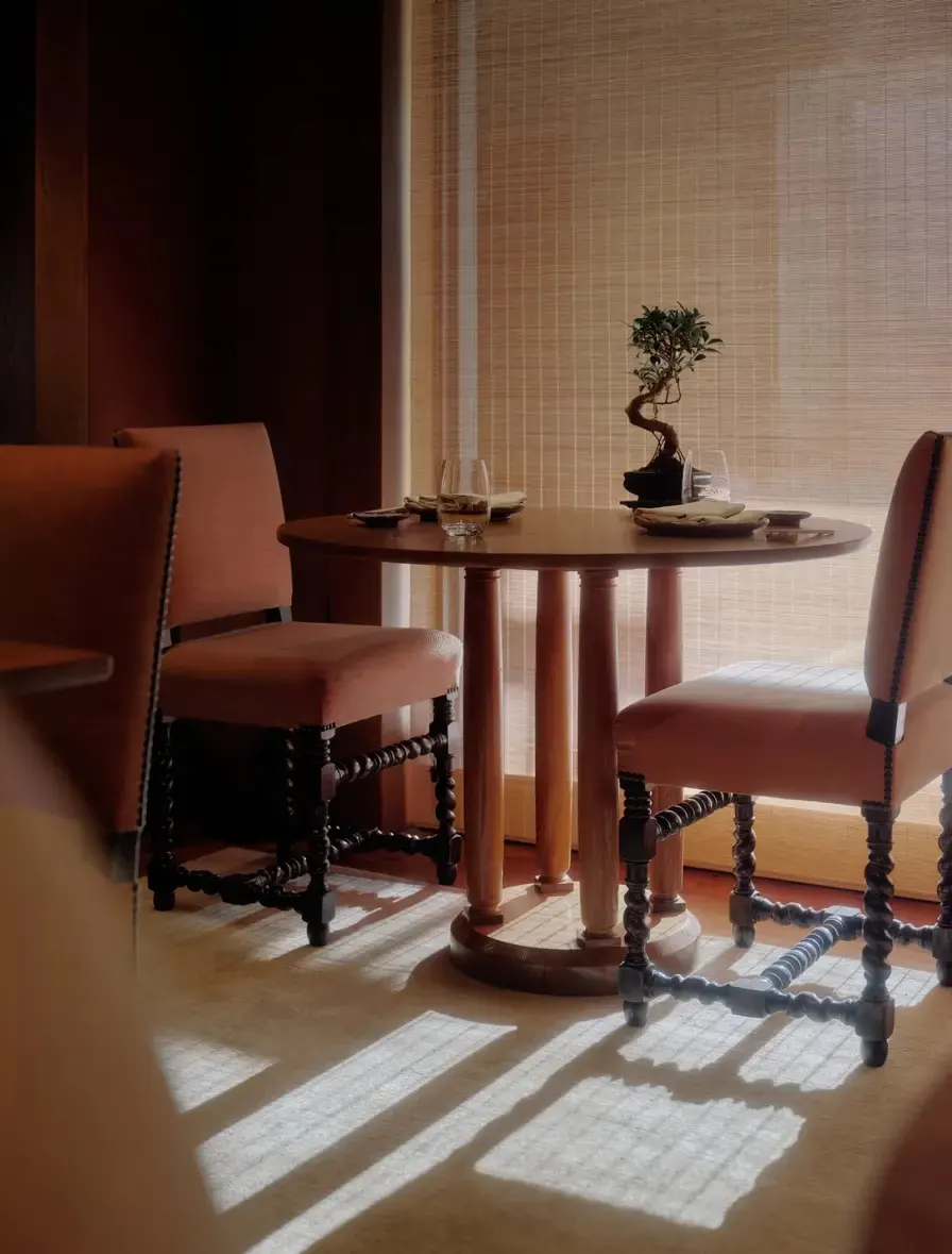 Table set for two with a bonsai centerpiece, terracotta chairs, and sunlight through woven blinds