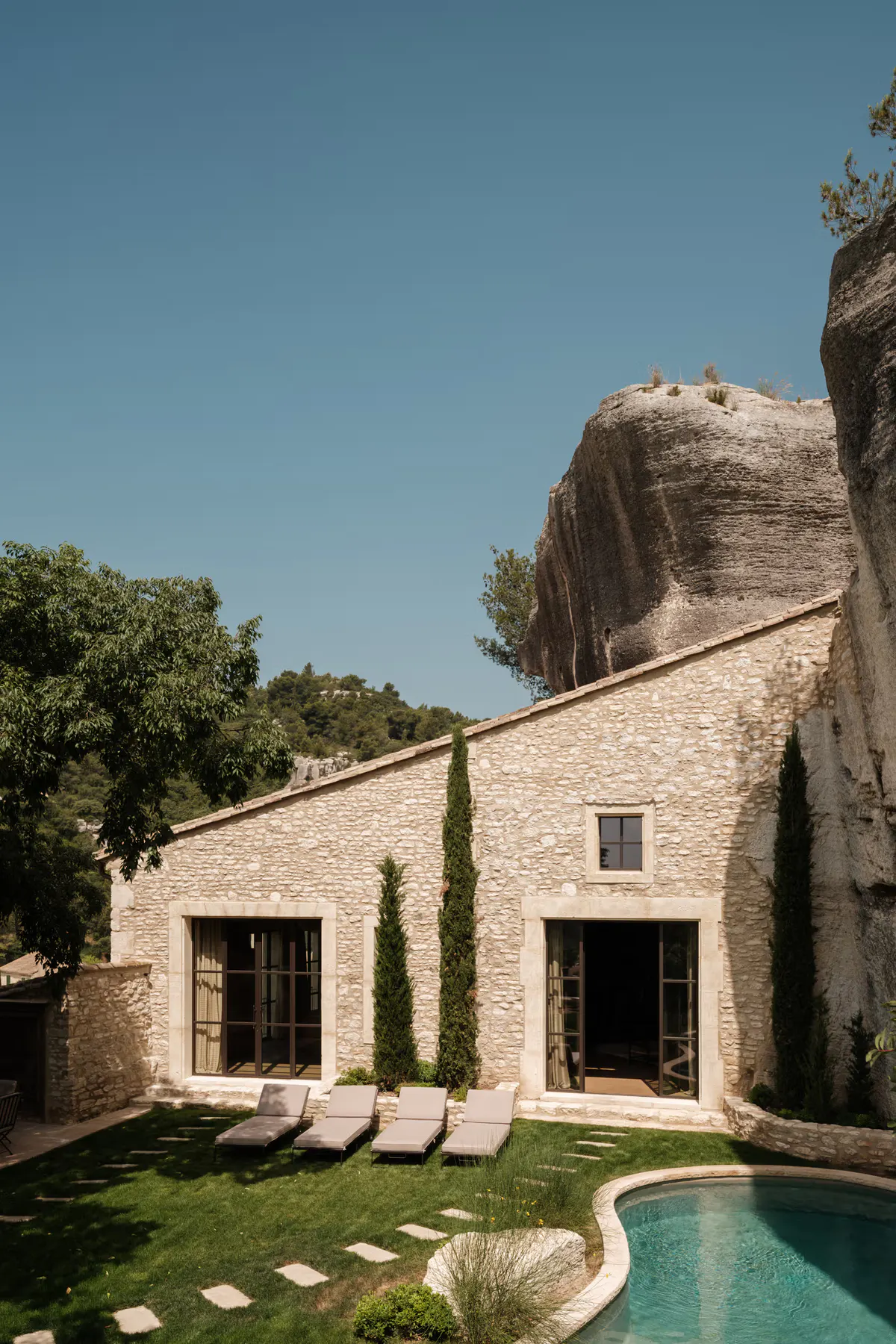 Stone courtyard with pale loungers, turquoise pool, raw limestone cliff wall, cypress wind-break privacy
