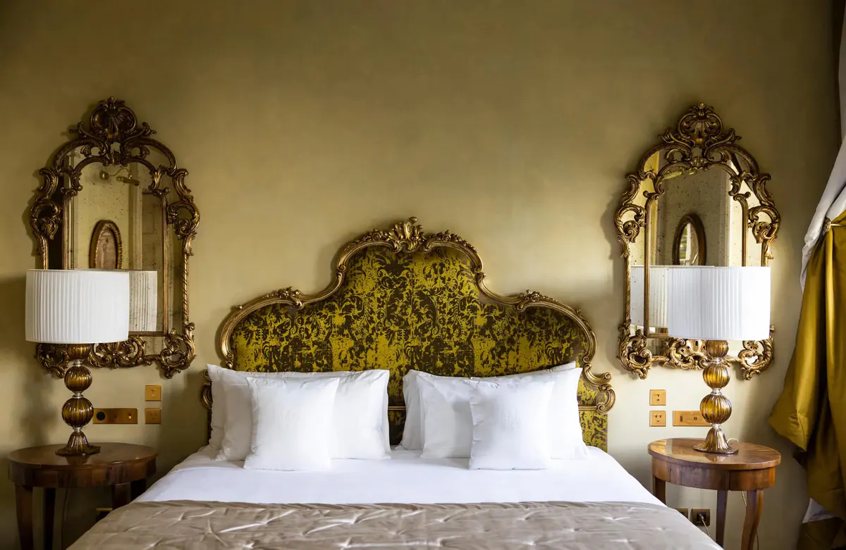 A bed with ornate gold headboard flanked by mirrored bedside tables and sconces