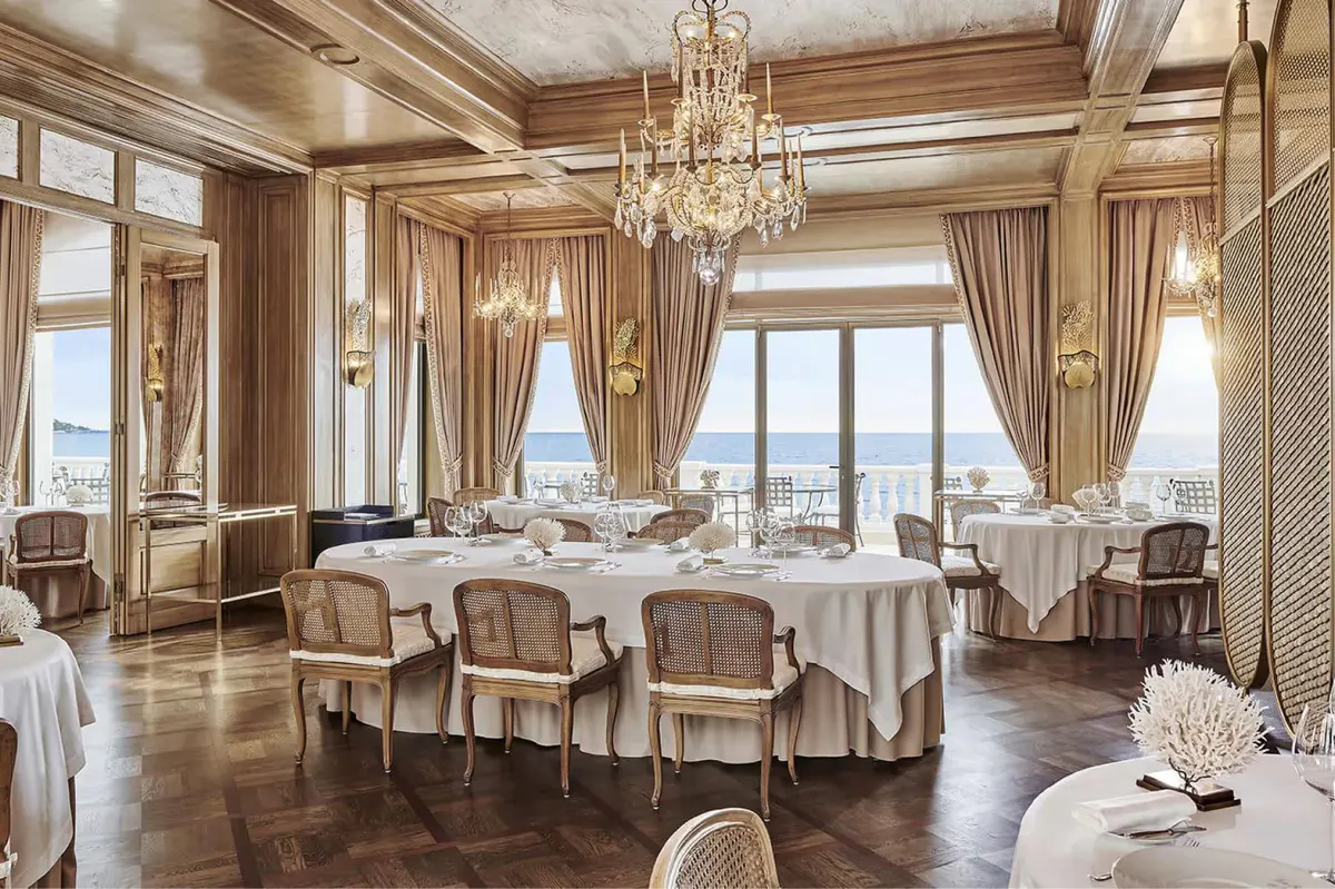 Grand salon dining space with round tables, high ceilings, and ocean backdrop