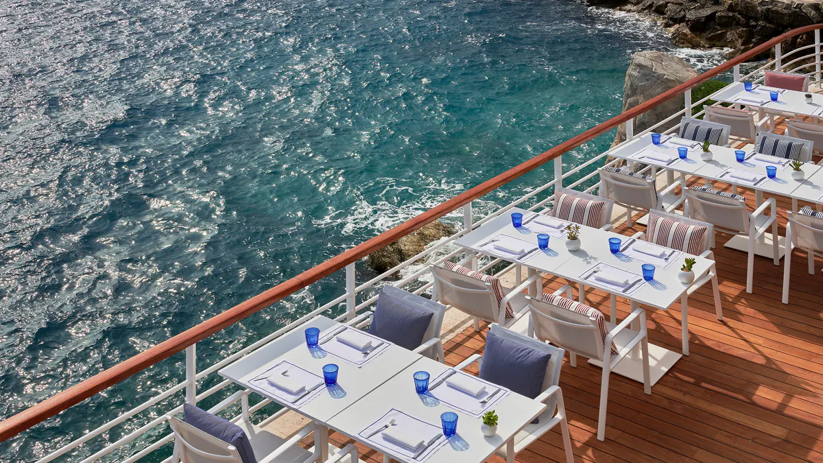 A seaside dining platform with white table settings, water views, and sailboats in the bay
