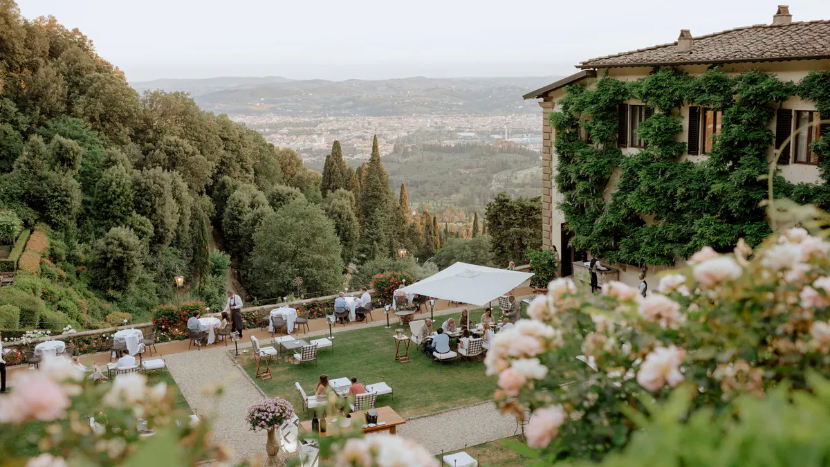 An afternoon garden party with guests at scattered tables, white umbrellas, and valley views beyond
