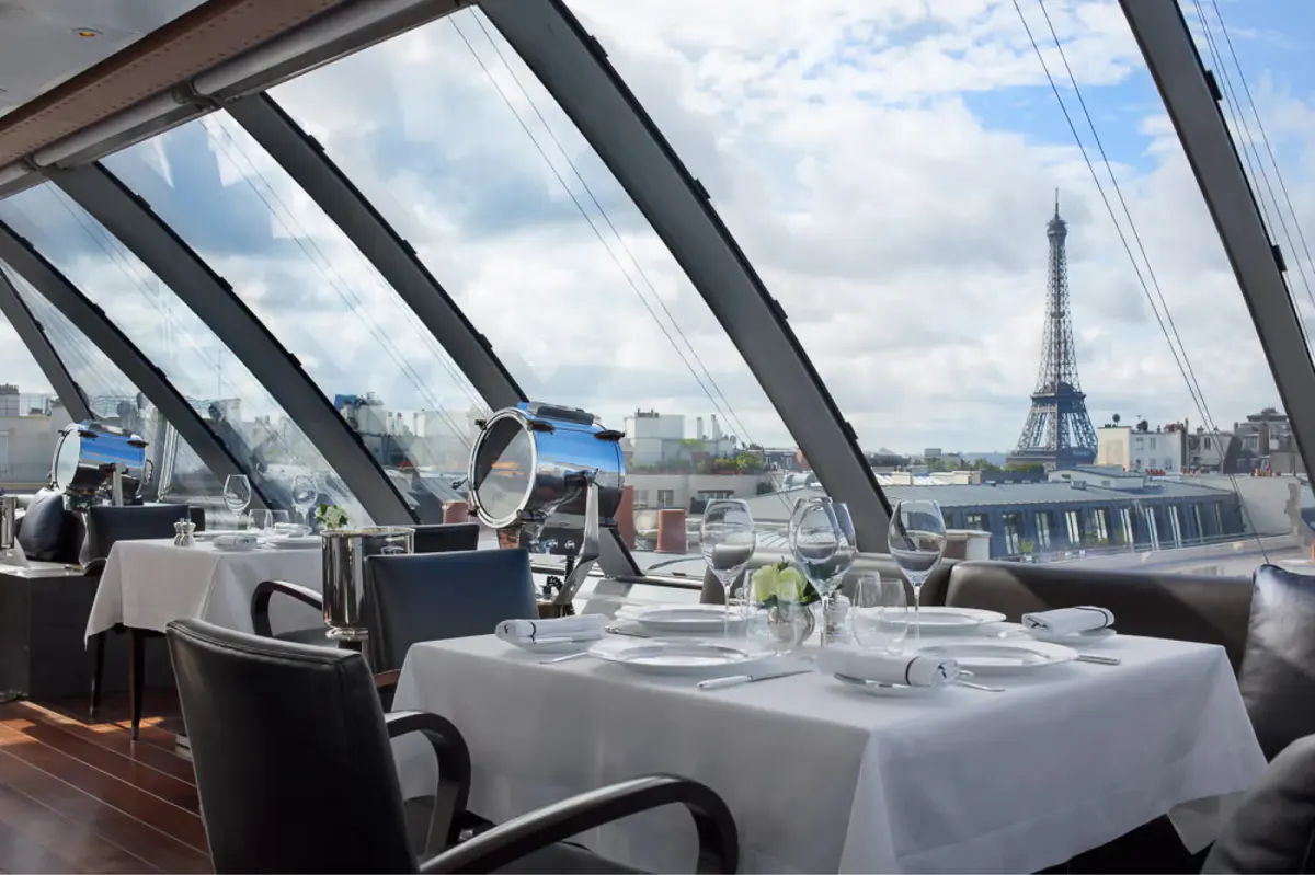 Rooftop dining terrace with glass panels, white tablecloths, and panoramic views of the Eiffel Tower