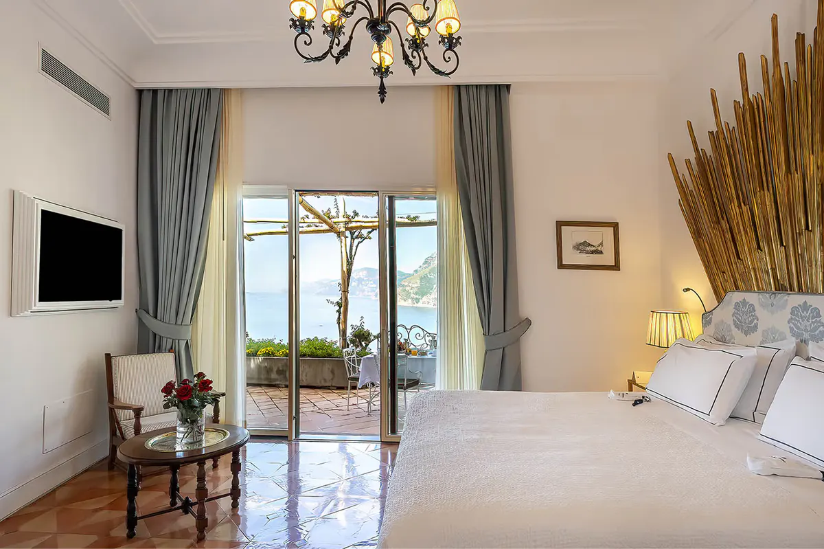 Bedroom suite with bed, chandelier, terrace doors, wood flooring, and glimpse of coastal landscape