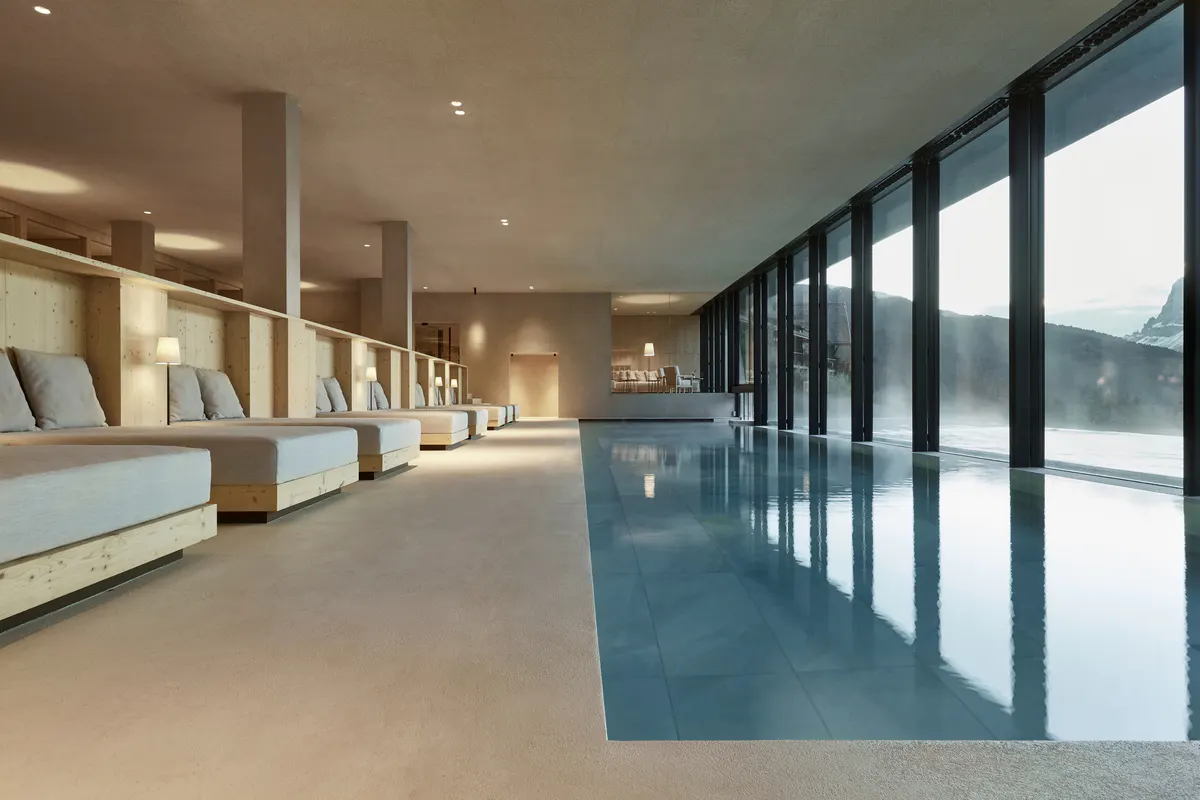 An indoor swimming pool with floor-to-ceiling glass walls overlooking mountains, light wood and stone finishes