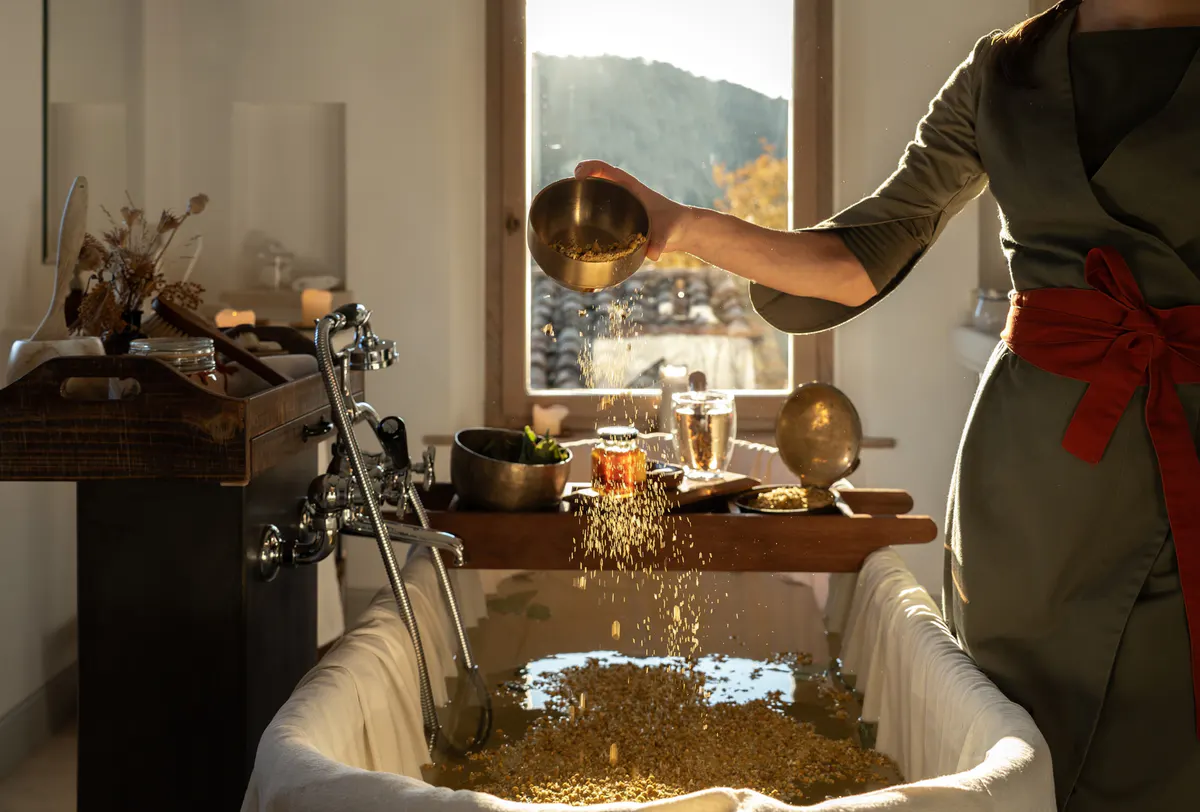 A robed attendant pouring bath salts into a golden vessel beside a window framing distant hills