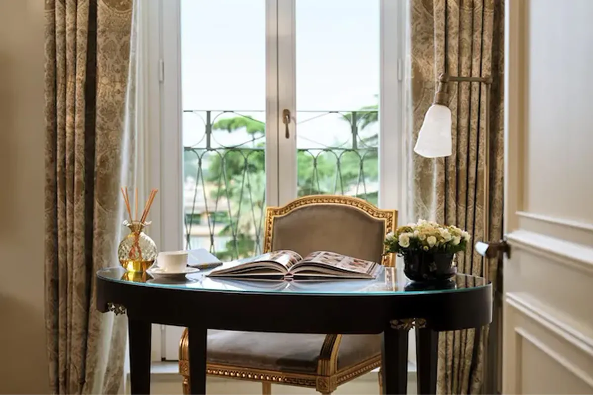 A writing desk by tall windows with gold curtains, topped with an open book and flowers overlooking the city