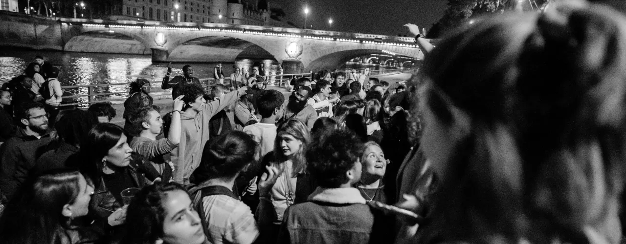 A night crowd celebrating on a Seine quai in Paris, a lit bridge behind, in black and white