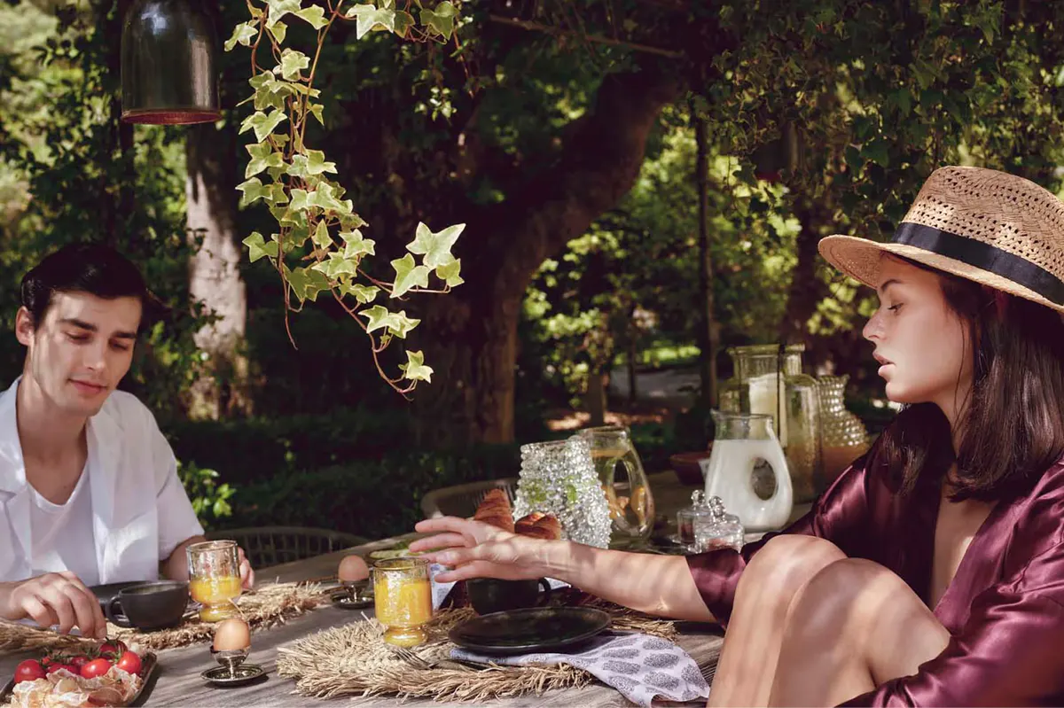 Two guests dining alfresco with candlelit table under dappled tree shade at dusk