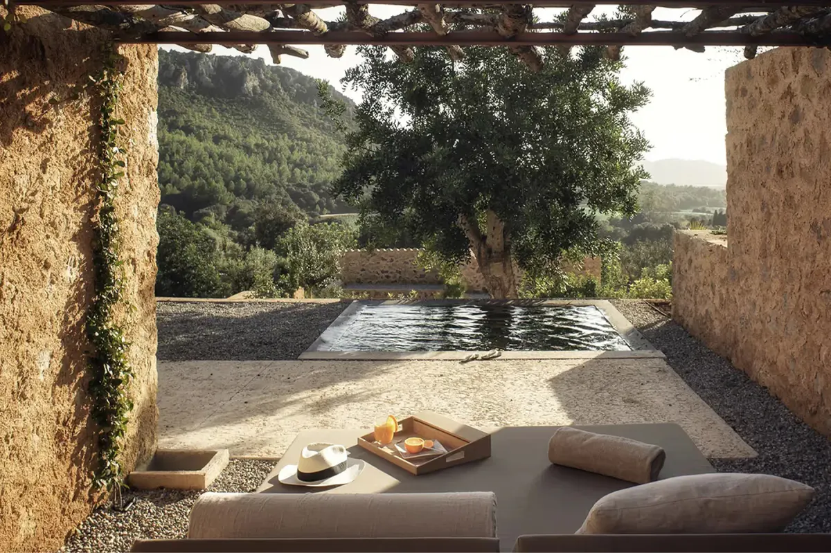 Covered lounge area framing a blue-toned pool, stone walls, and distant Mediterranean landscape views