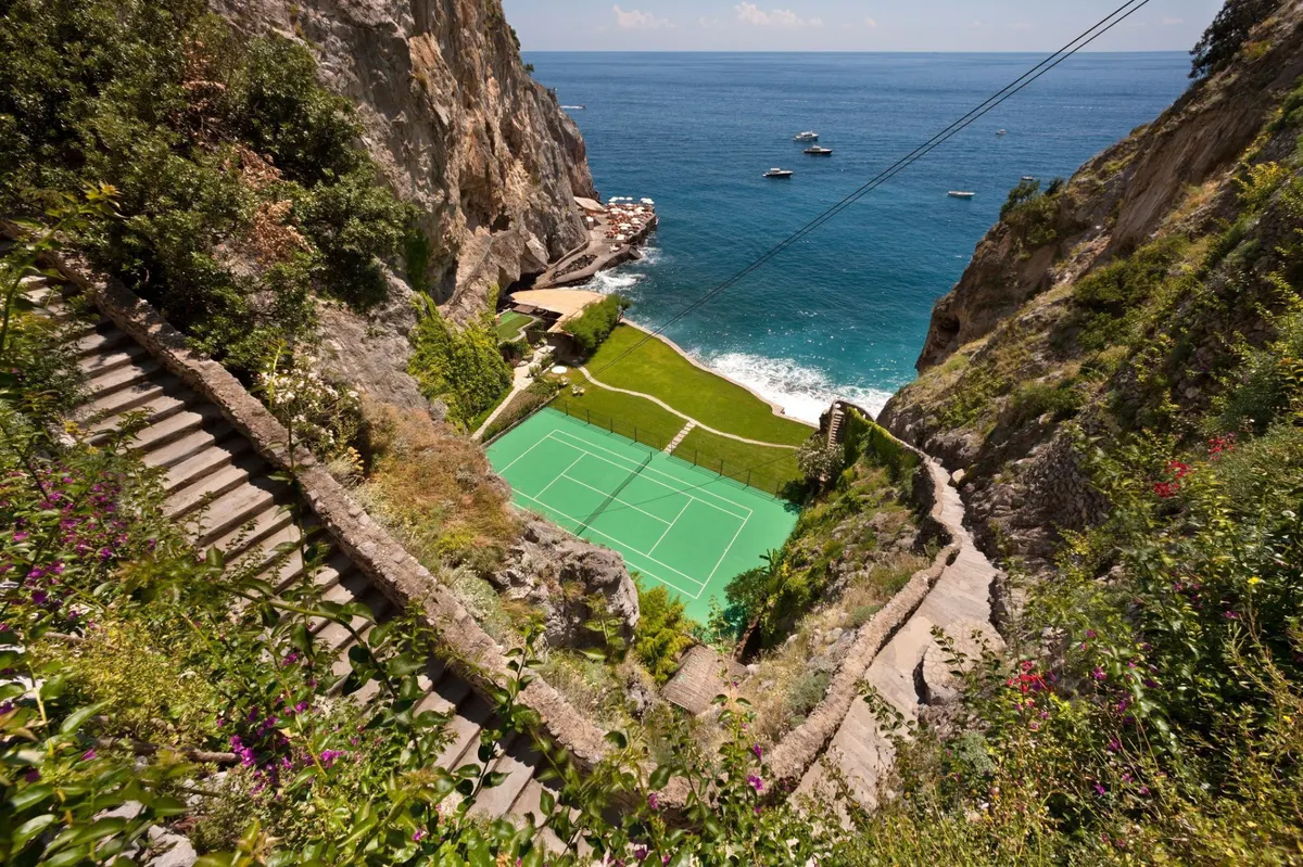 Green tennis court carved into cliffside between green vegetation and bright Mediterranean sea