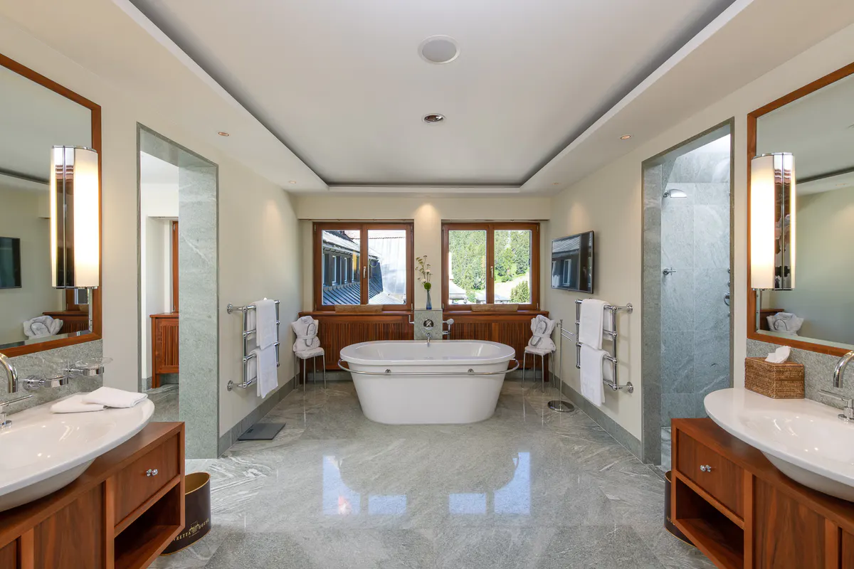 A spa bathroom with dual sinks, a soaking tub, large windows, and neutral stone finishes