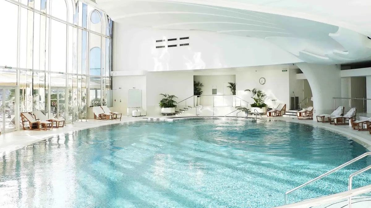Indoor swimming pool with natural light and rows of lounge seating along deck