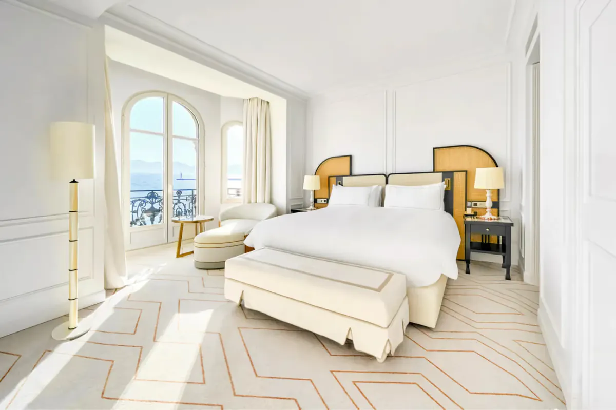 Bedroom with white bed, brass-trimmed nightstands, geometric floor pattern, and glass doors to a balcony with sea views