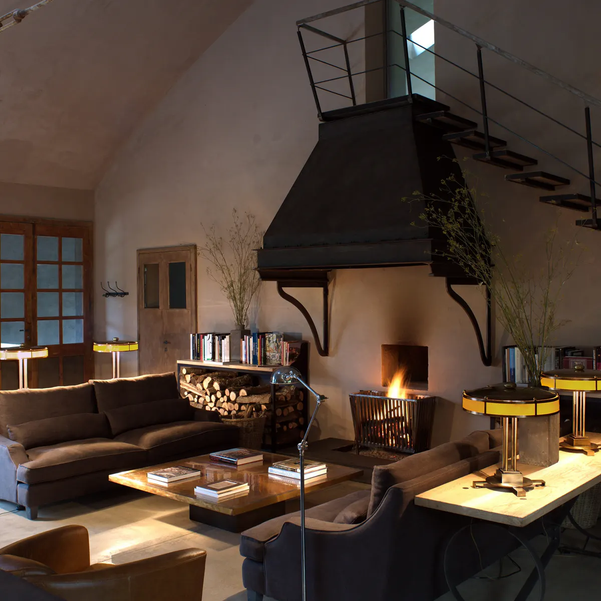 A double height living room with a black hooded fireplace, leather sofas, and a mezzanine loft above