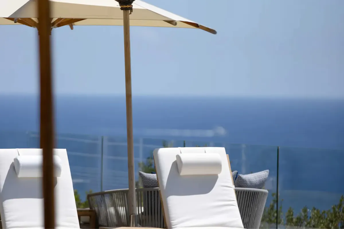 Two white lounges beneath a wooden umbrella with Mediterranean horizon