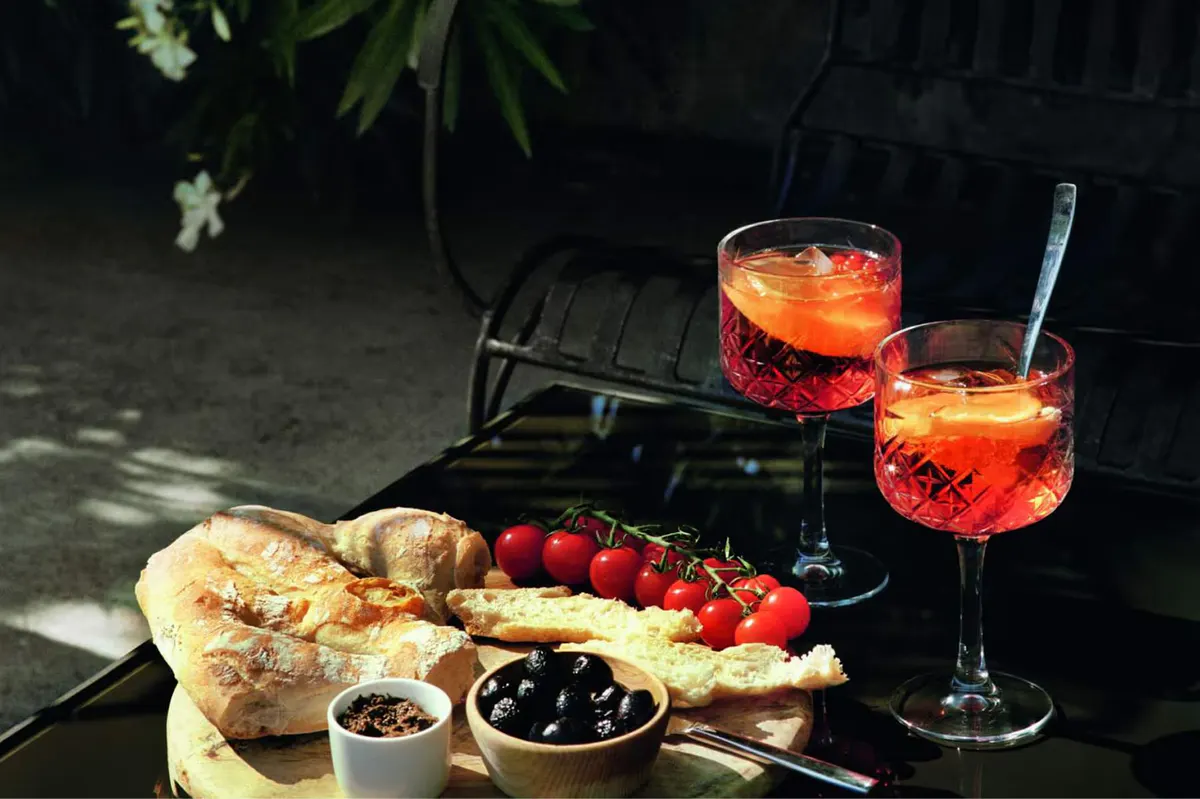 Aperitif with fresh bread, olives, and red wine on outdoor table with greenery backdrop