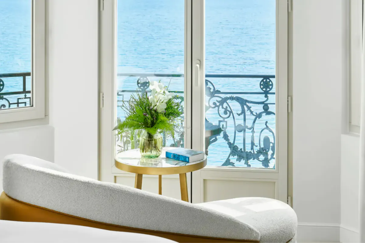 Window nook with white upholstered seating overlooking the sea, with white-painted French doors and a tall plant