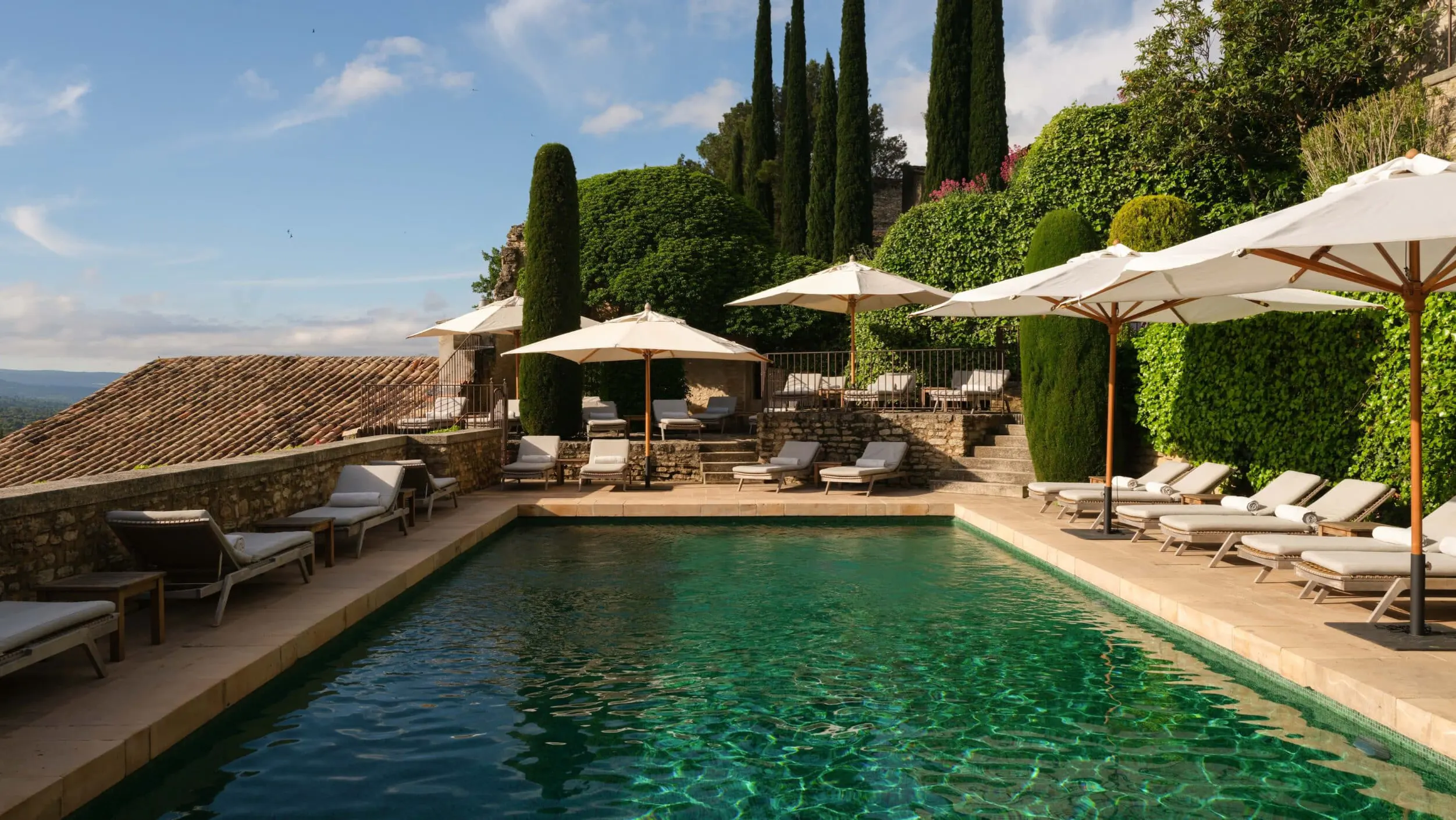 A swimming pool overlooking rolling vineyards and mountains under a blue sky with cypress trees and lounging areas