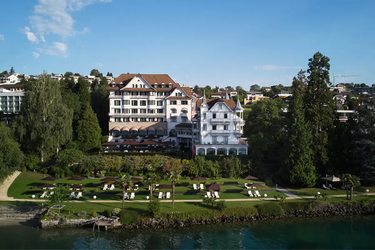 Whitewashed hotel building with turret beside Lake Lucerne, surrounded by mature trees
