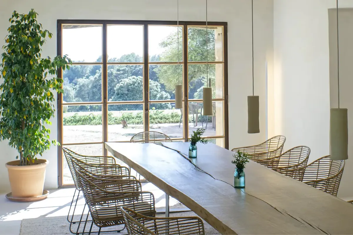 Contemporary dining room with long table, woven chairs, and full-height windows overlooking the estate