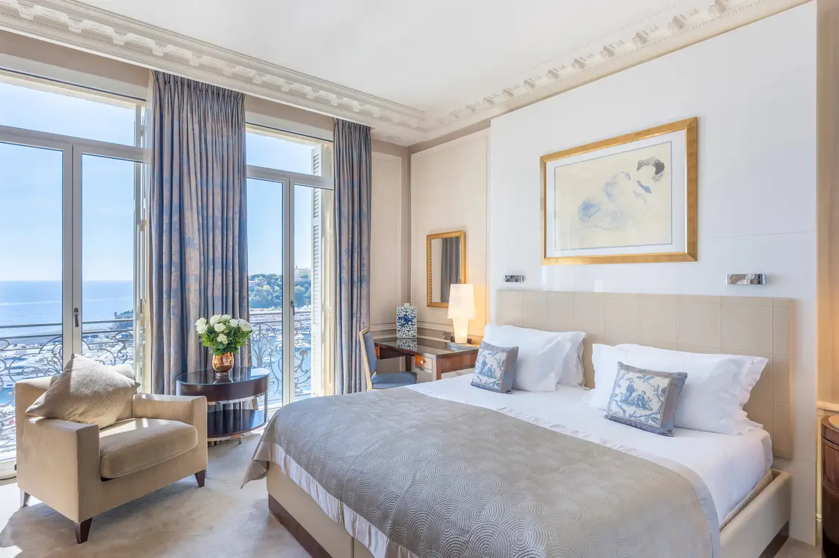 Master bedroom with white duvet, gold fixtures and chair seating near arched terrace
