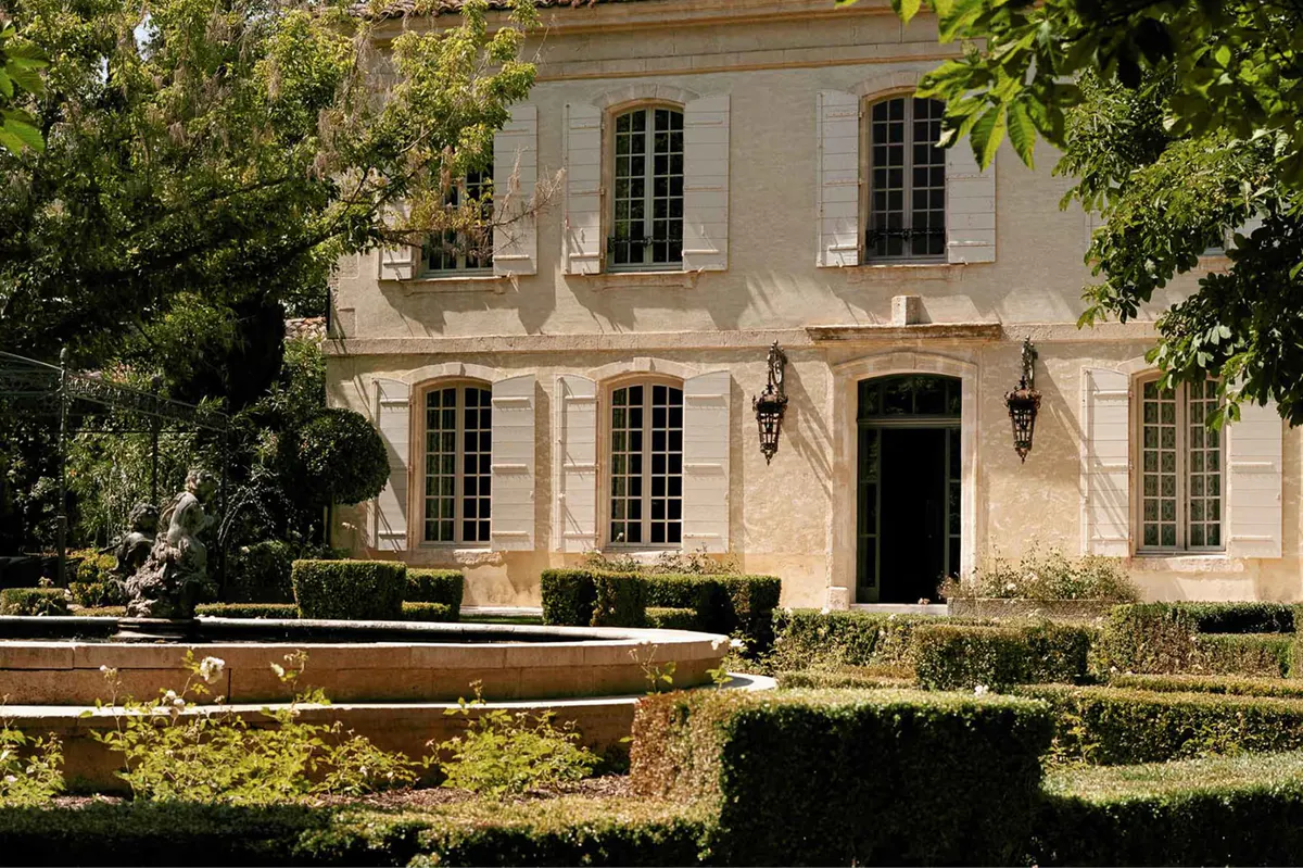 Pale stone facade with arched entry, dark shutters, and manicured hedge border
