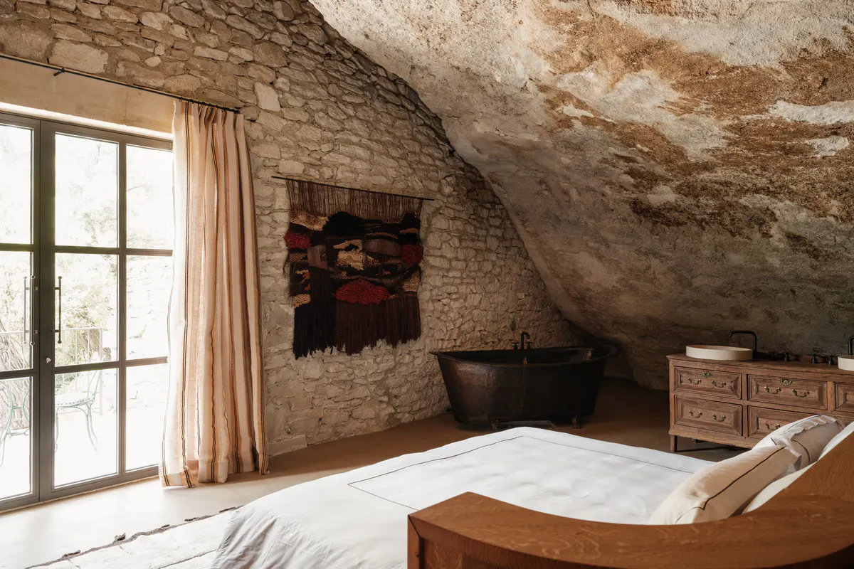 Rock-carved bedroom with curved limestone walls, black cast-iron soaking tub, wooden dresser, Berber textile art