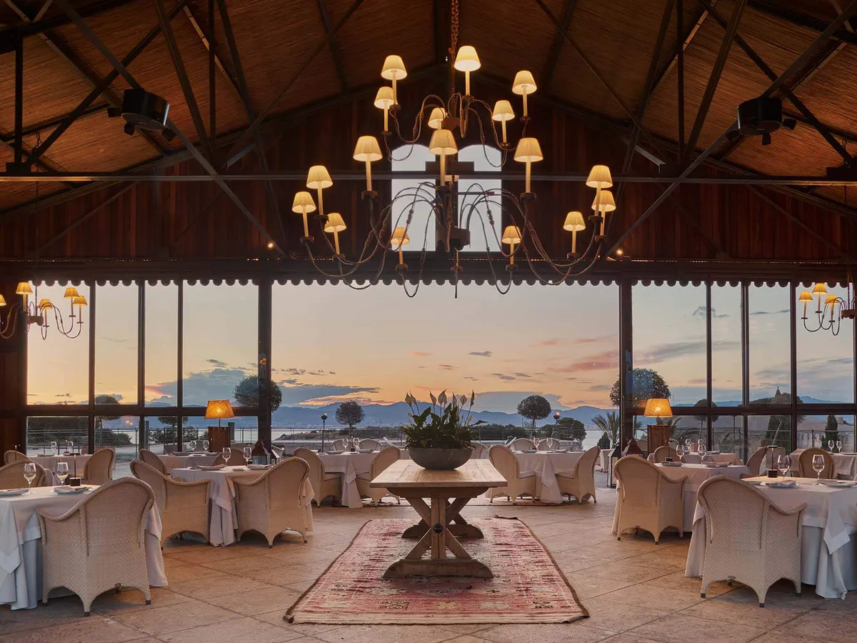 A vast dining pavilion with wooden beam trusses, brass lanterns, cream chairs, and panoramic sea views