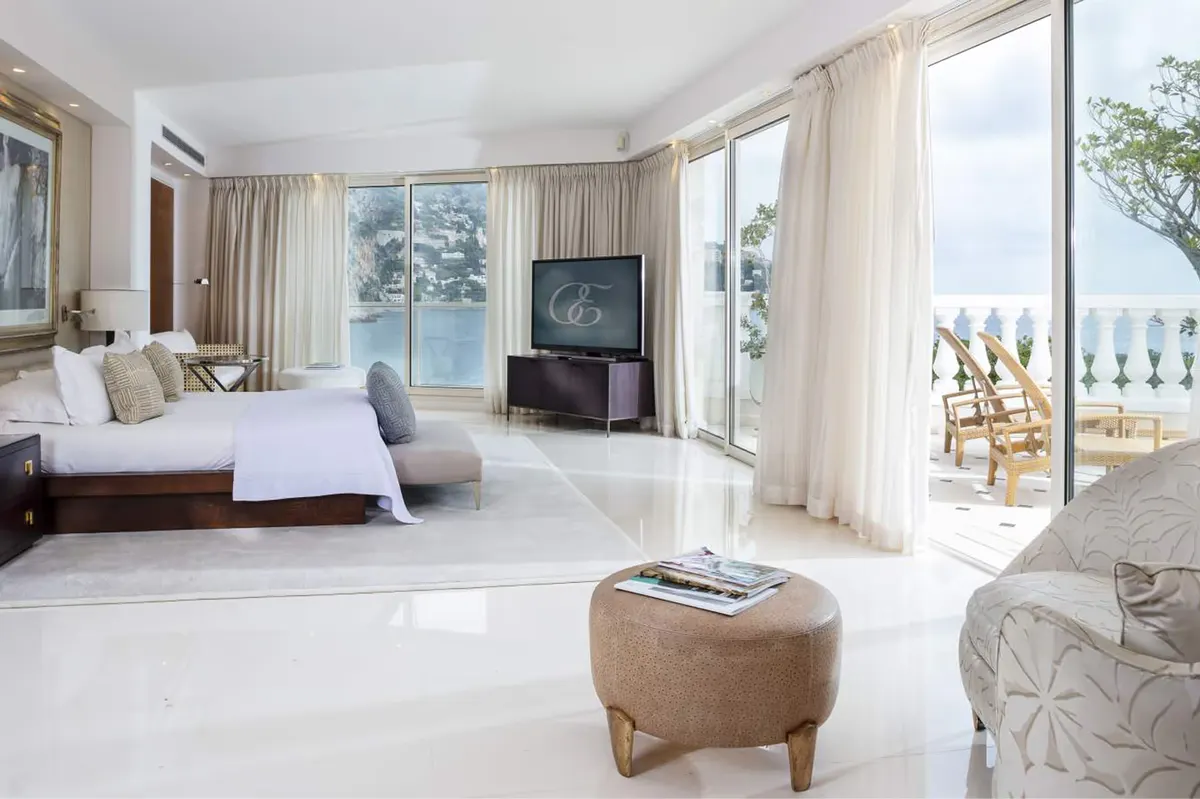 A bright bedroom with white furnishings, sheer curtains, and glass doors opening to sea views