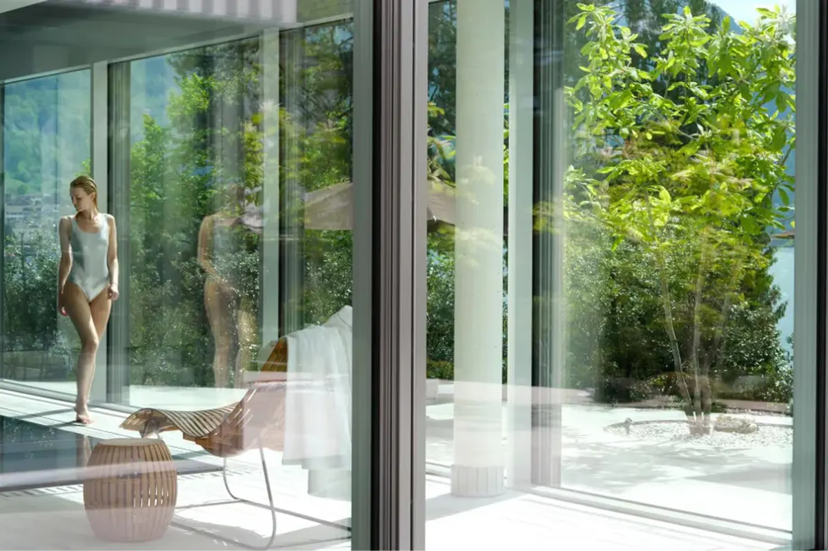 Indoor spa pool area with water view, woman in spa dress on white lounger by glass wall