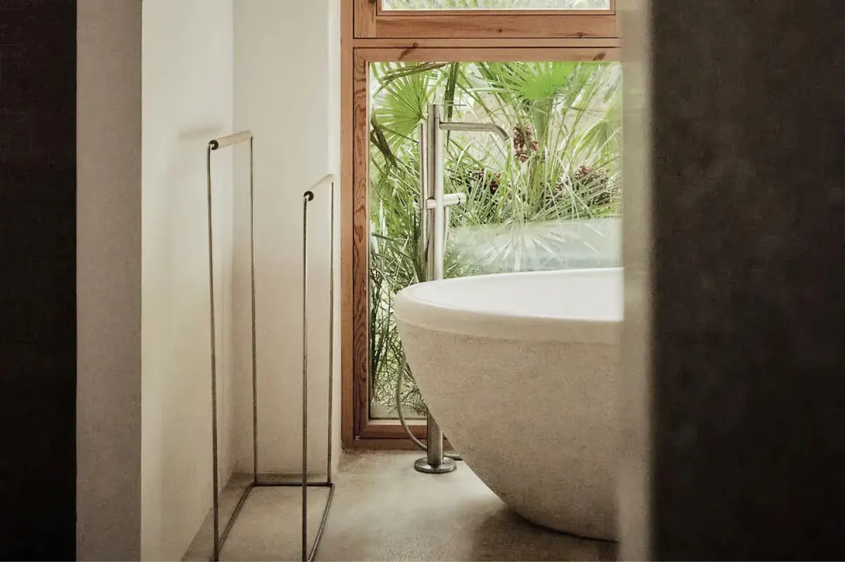 Freestanding sculptural bathtub positioned beside metal-frame window overlooking verdant landscape