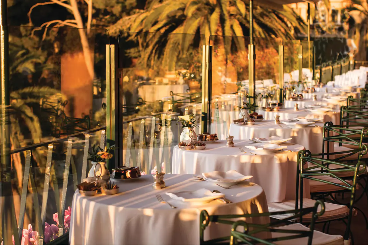 An outdoor dining terrace with white tablecloths, tropical plants, and sea backdrop