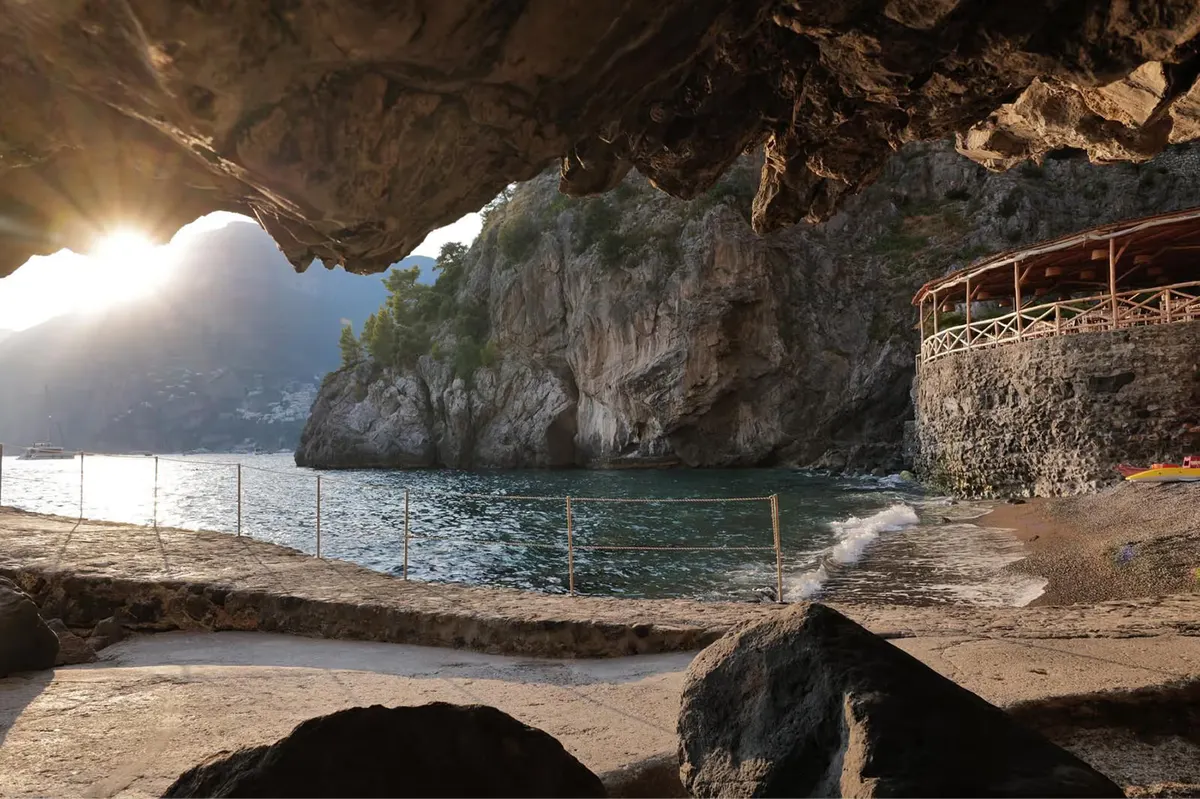 Private beach cove beneath limestone arch, sun-flooded cave opening, calm water, safety railings, wooden dock