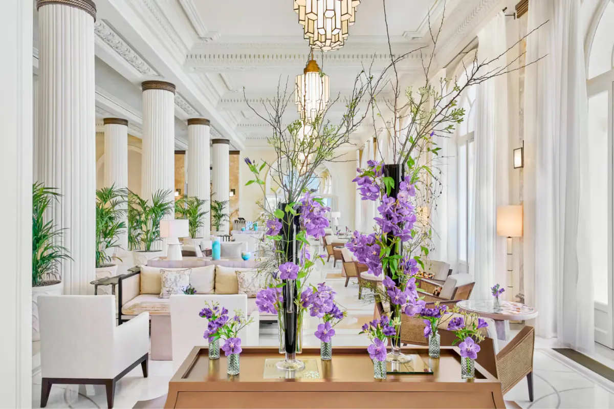 Bright lobby with tall white columns, purple flower arrangements on a wooden table, and chandelier lighting