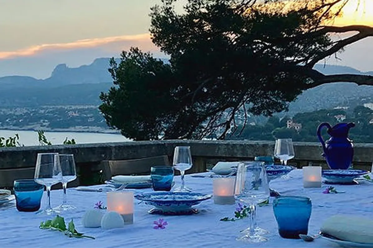 Outdoor dining setup at dusk overlooking turquoise sea with coastal landscape backdrop