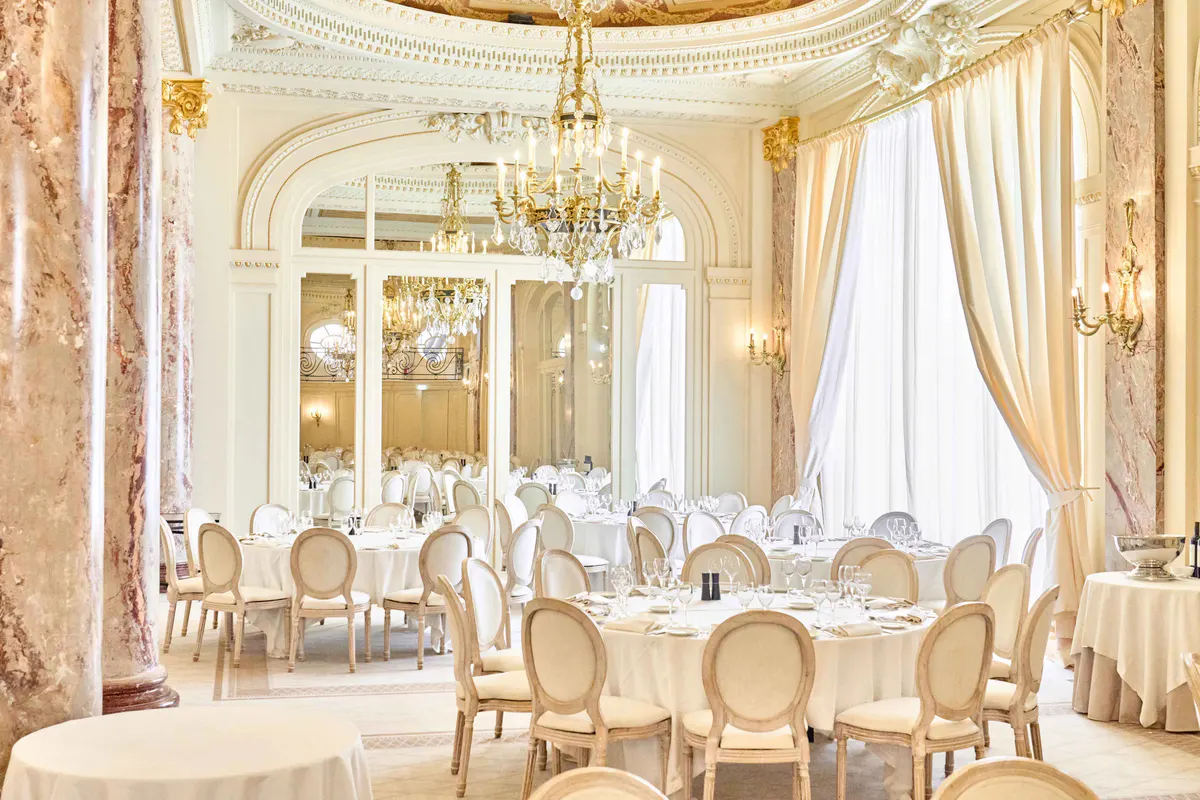 Elegant dining room with gilt trim, cream-colored walls, white tablecloths, and pale wood chairs under a crystal chandelier