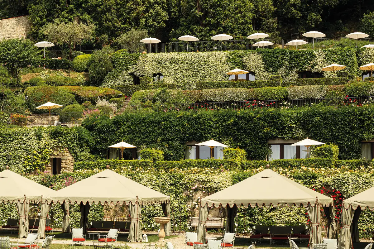 Loungers beneath striped umbrellas in a terraced garden with flowering plants and cypress trees